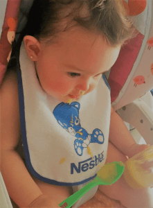 Victoria comiendo sola con su babero de Nestlé y su cuchara de Ordesa