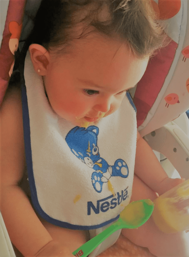 Victoria comiendo sola con su babero de Nestlé y su cuchara de Ordesa